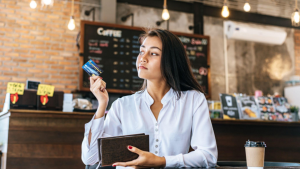 A imagem mostra uma mulher jovem sentada em uma cafeteria moderna, segurando um cartão de crédito e uma carteira enquanto olha para o lado com uma expressão pensativa.