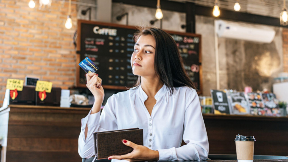 A imagem mostra uma mulher jovem sentada em uma cafeteria moderna, segurando um cart&atilde;o de cr&eacute;dito e uma carteira enquanto olha para o lado com uma express&atilde;o pensativa.