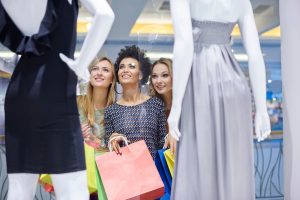 Tr&ecirc;s mulheres animadas fazendo compras e admirando a vitrine de loja, encantadas com os vestidos em exposi&ccedil;&atilde;o.