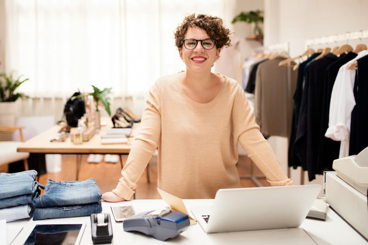 Mulher sorridente em ambiente de loja, rodeada por roupas e produtos diversos, representando a atividade de vendas e atendimento ao cliente.