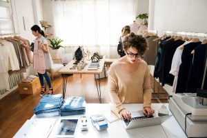 Mulher em loja de roupas organizando vendas no laptop, representando como escolher o melhor ponto comercial para loja de roupas.