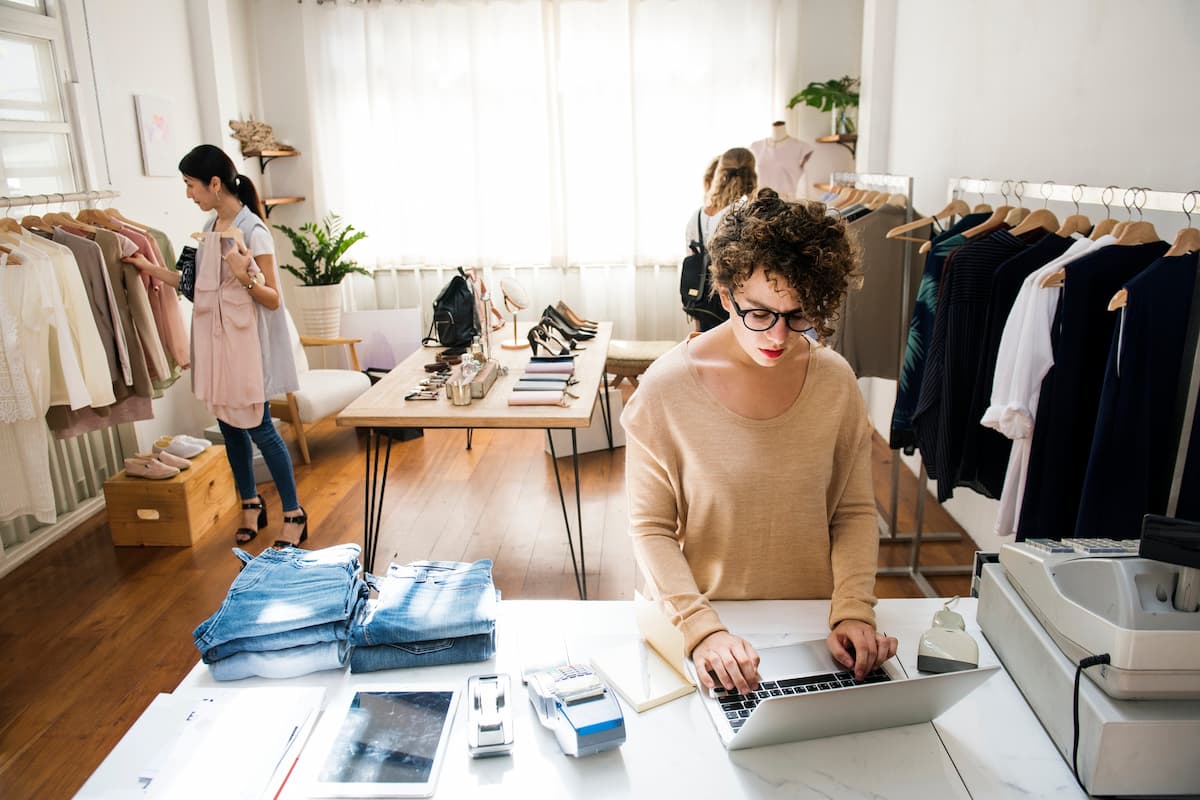 Mulher em loja de roupas organizando vendas no laptop, representando como escolher o melhor ponto comercial para loja de roupas.