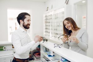 Homem e mulher sorrindo enquanto experimentam óculos em uma franquia de ótica moderna e bem iluminada.