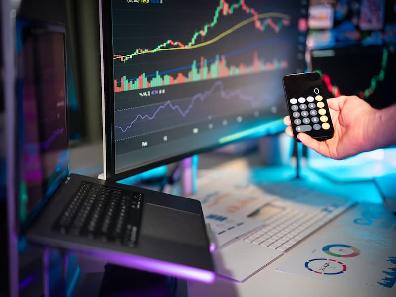 A imagem mostra um ambiente de trabalho com múltiplas telas exibindo gráficos financeiros, uma pessoa segurando um smartphone com a calculadora aberta, além de documentos e gráficos em papel na mesa. A cena remete diretamente a um cenário de análise de dados, finanças e tomada de decisões.