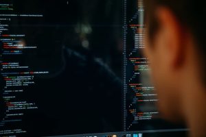 A imagem mostra uma pessoa observando atentamente uma tela de computador com código-fonte — provavelmente relacionado a desenvolvimento de software. O código parece estar escrito em JavaScript (ou JSX), comum em aplicações web. Essa cena sugere um ambiente de desenvolvimento técnico voltado à criação de soluções digitais, como ferramentas de análise de crédito automatizadas.