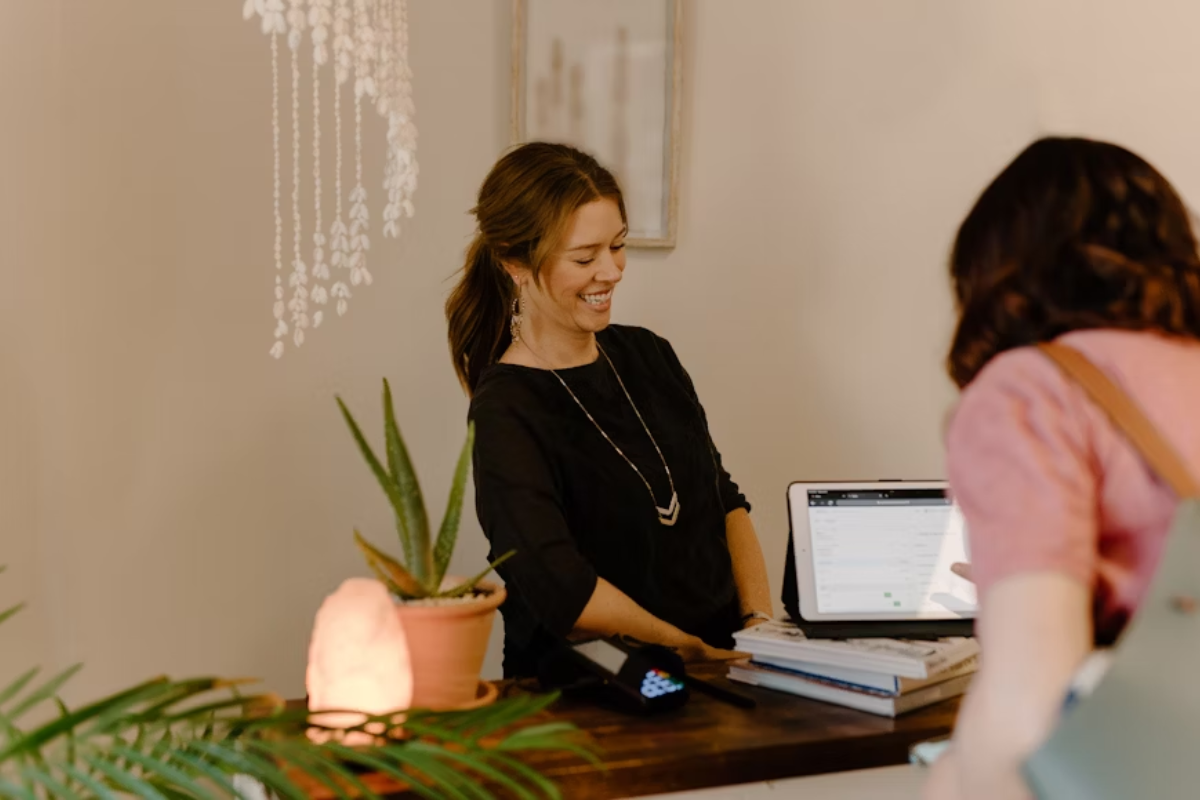 Uma funcionária sorri enquanto atende uma cliente em um ambiente de loja física. Há uma atmosfera amigável e próxima, com elementos decorativos como plantas, uma luminária de sal e livros sobre o balcão.