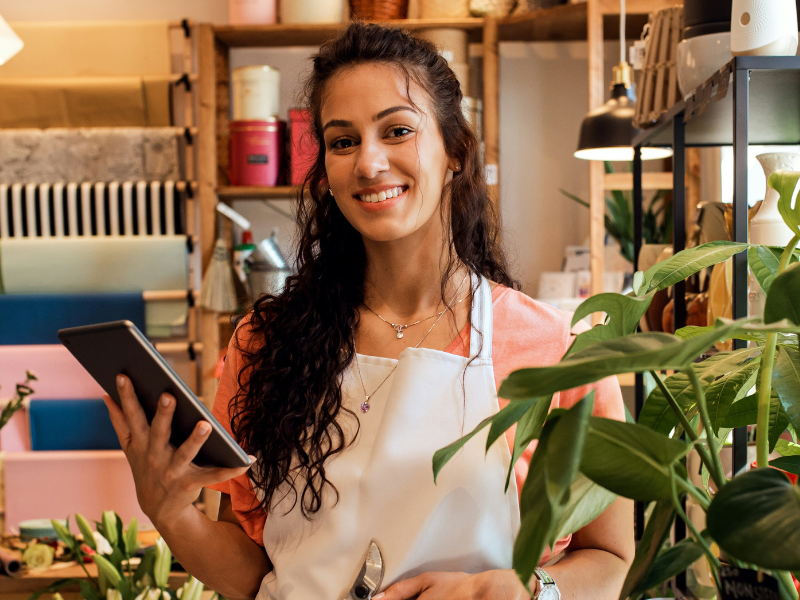 A imagem mostra uma jovem mulher de cabelos castanhos longos e cacheados, com um sorriso amigável, em um ambiente de loja. Ela parece ser a dona ou funcionária do local.