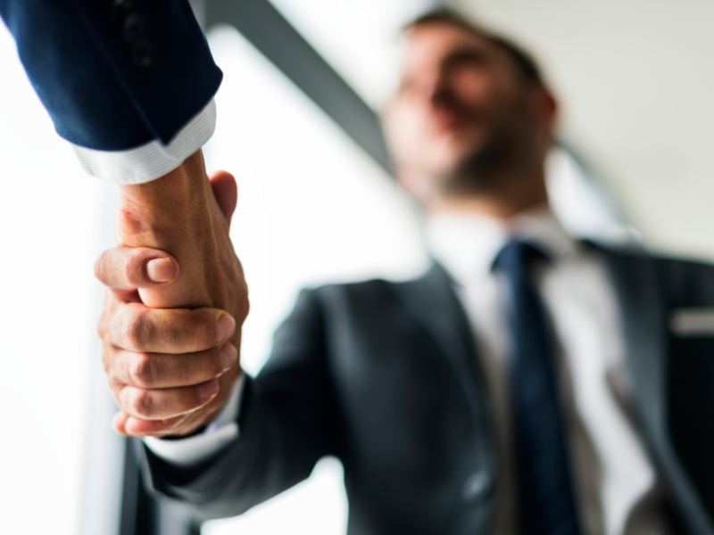 A imagem mostra um aperto de m&atilde;o entre duas pessoas, com o foco principal sendo as m&atilde;os e o gesto de cumprimento. Uma das pessoas est&aacute; usando um terno escuro com camisa branca e gravata, transmitindo uma apar&ecirc;ncia formal e corporativa. A outra pessoa, cujo rosto n&atilde;o &eacute; vis&iacute;vel, tamb&eacute;m parece estar vestida formalmente.