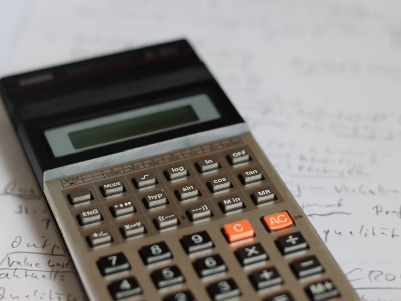 A imagem &eacute; um close-up (plano detalhe) de uma calculadora cient&iacute;fica ou financeira de modelo antigo, sobreposta a um fundo de papel manuscrito.