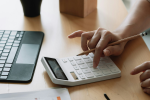A imagem mostra uma pessoa usando uma calculadora branca sobre uma mesa de madeira clara. A mão direita pressiona as teclas da calculadora enquanto segura um lápis, sugerindo que está realizando cálculos ou análises financeiras.