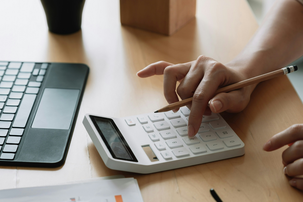 A imagem mostra uma pessoa usando uma calculadora branca sobre uma mesa de madeira clara. A m&atilde;o direita pressiona as teclas da calculadora enquanto segura um l&aacute;pis, sugerindo que est&aacute; realizando c&aacute;lculos ou an&aacute;lises financeiras.