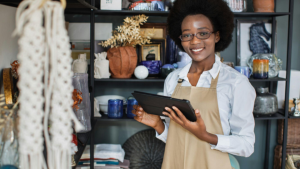 Esta imagem captura uma mulher em pé e sorrindo, em um espaço de trabalho. Ela está usando óculos, uma camisa de manga comprida de cor clara e um avental de chef, o que sugere que ela está trabalhando. Ela tem um sorriso no rosto e olha para você com seus olhos expressivos.