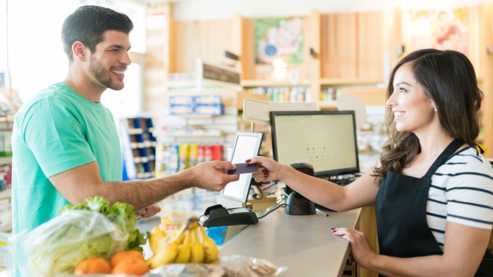Esta imagem &eacute; um plano m&eacute;dio de um cliente do sexo masculino entregando um cart&atilde;o de cr&eacute;dito a uma caixa do sexo feminino em um caixa de supermercado.