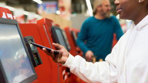 A imagem mostra uma pessoa utilizando um terminal de autoatendimento em um ambiente que parece ser um supermercado, loja ou estação de serviços automatizados.