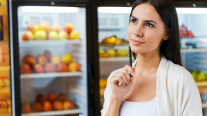 A imagem mostra uma mulher em frente a uma geladeira aberta, aparentemente em um momento de decisão. Ela está segurando uma caneta próxima ao rosto, com uma expressão pensativa, olhando levemente para cima, como se estivesse refletindo sobre o que escolher.